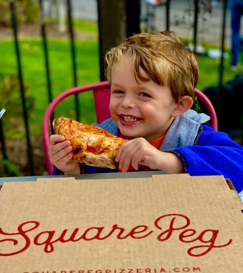 A happy kid eating a slice of Square Peg pizza